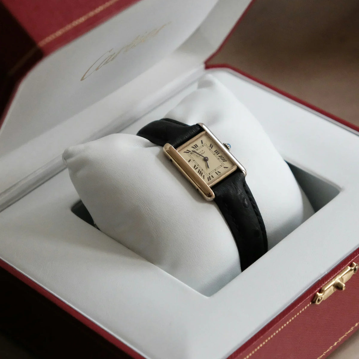 Cartier Tank watch with black leather strap and gold-tone case, resting on its cushion inside an open red Cartier presentation box.