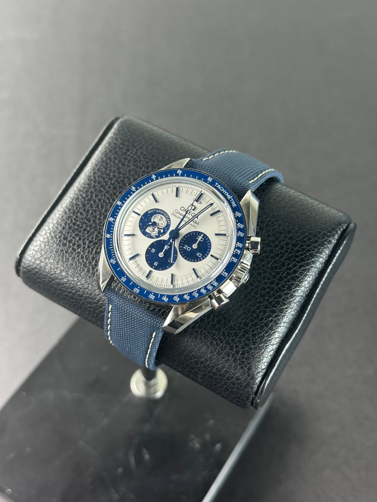 Montre Omega Speedmaster Professional Silver Snoopy Award 50e anniversaire à cadran argenté et bracelet tissu bleu, posée sur un coussin en cuir noir